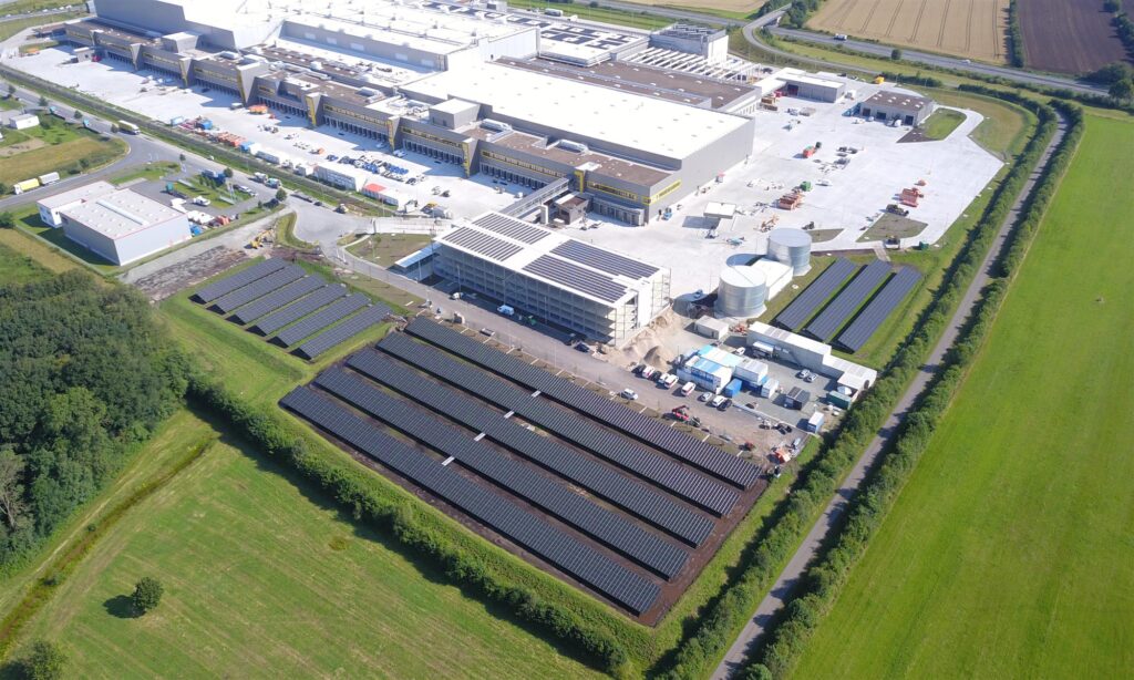 Drohnenaufnahme des Solarparks Neumünster mit Photovoltaik-Modulreihen neben einem Industriekomplex